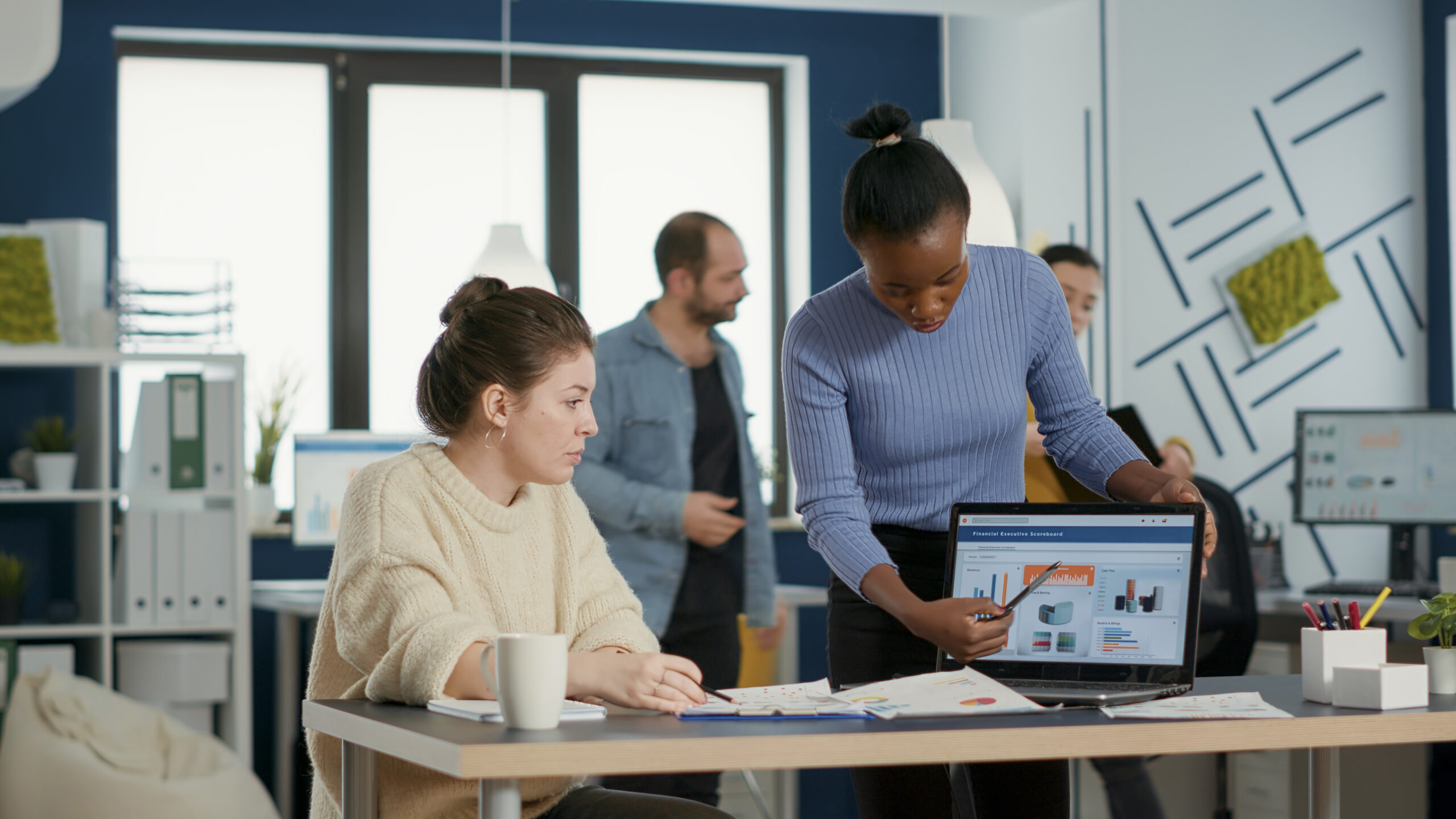 African american startup employee presenting marketing strategy on laptop to coworker drinking cofee or tea from cup. Casual colleagues working on group project analyzing business annual turnover.